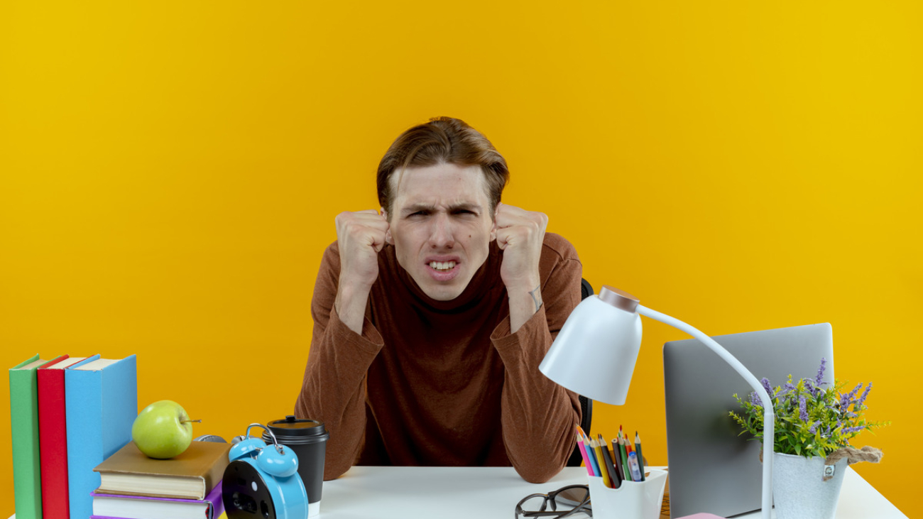 concerned-young-student-boy-sitting-desk-with-school-tools-showing-yes-gesture (1)